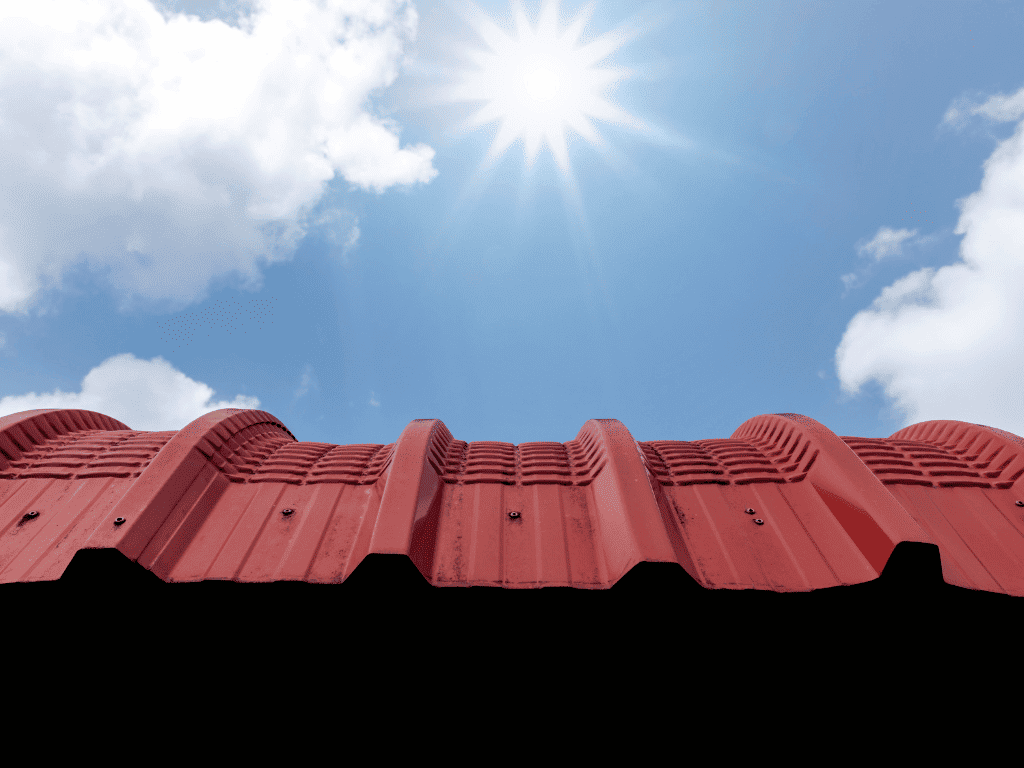Upward view of a red corrugated metal roof under a sunny sky with scattered clouds and visible sun rays.